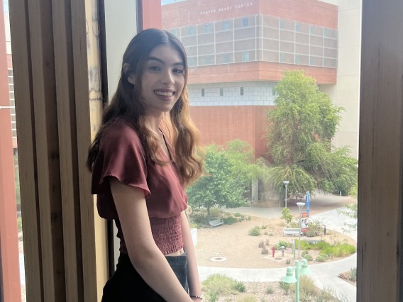 photograph of a person standing next to a window with the University of Arizona campus in the background